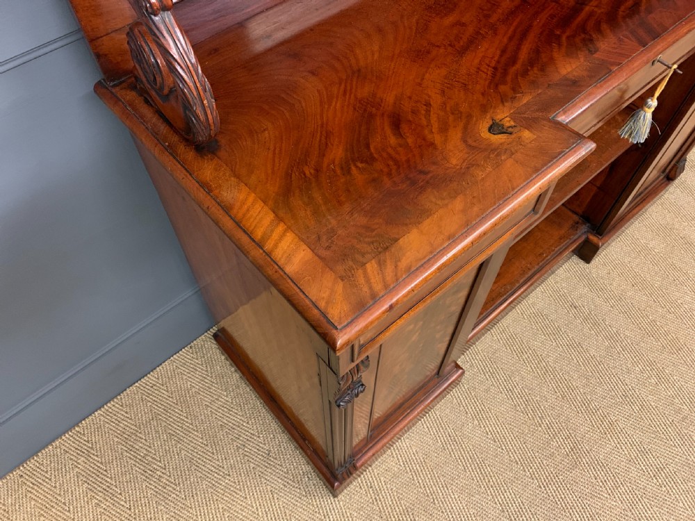 An early nineteenth-century flame mahogany chiffonier featuring a carved ornamental cresting, open central bookshelves, and two side cupboards. View VIII