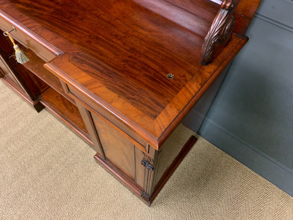 An early nineteenth-century flame mahogany chiffonier featuring a carved ornamental cresting, open central bookshelves, and two side cupboards. View IX