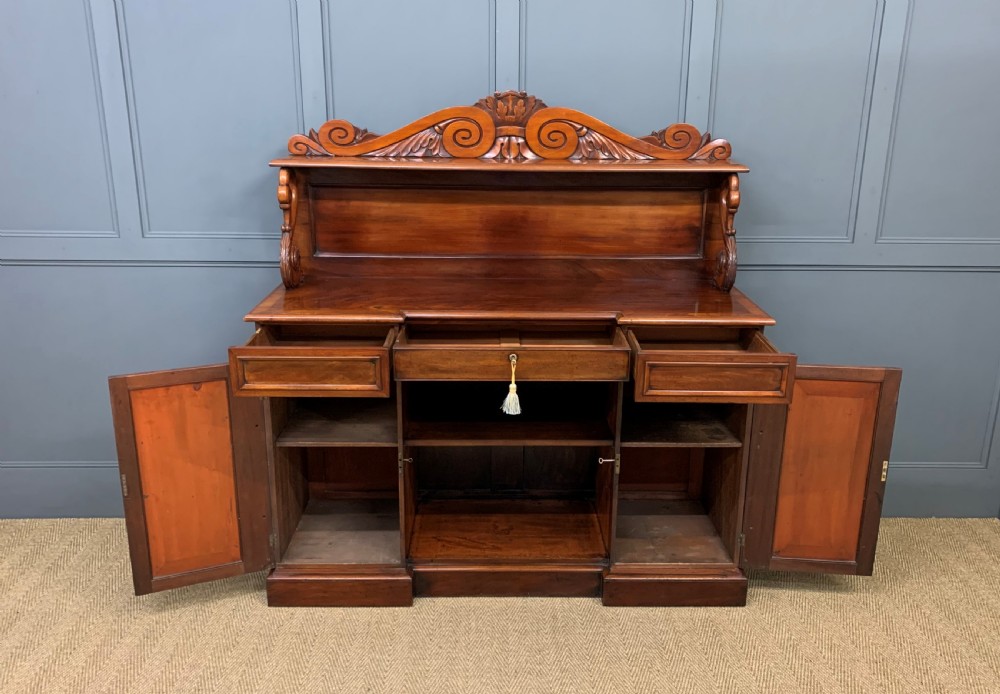 An early nineteenth-century flame mahogany chiffonier featuring a carved ornamental cresting, open central bookshelves, and two side cupboards. View XIII