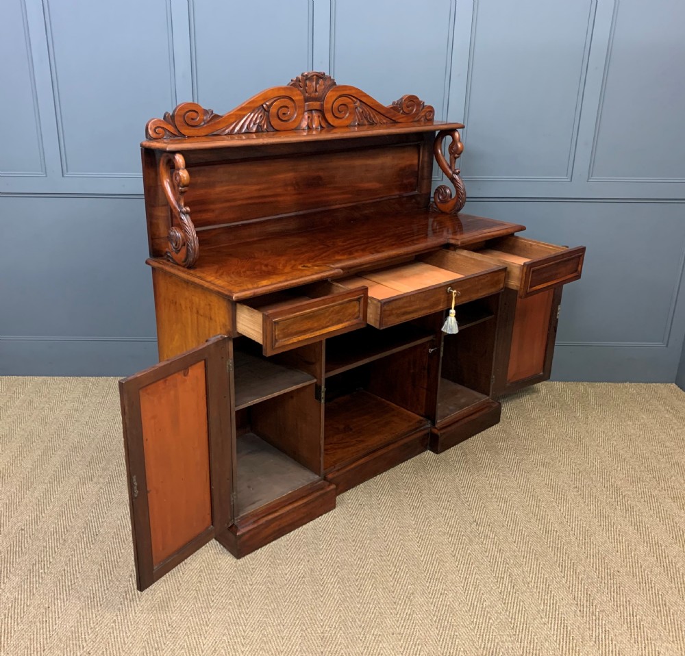 An early nineteenth-century flame mahogany chiffonier featuring a carved ornamental cresting, open central bookshelves, and two side cupboards. View XVIII