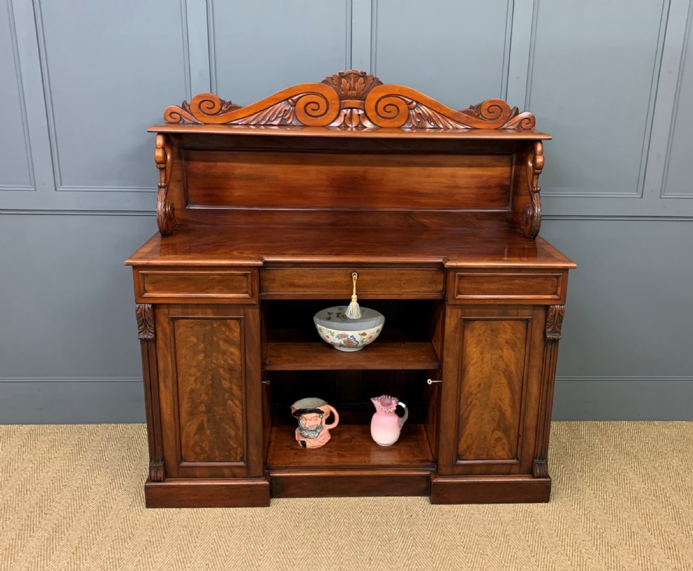 An early nineteenth-century flame mahogany chiffonier featuring a carved ornamental cresting, open central bookshelves, and two side cupboards. View XX