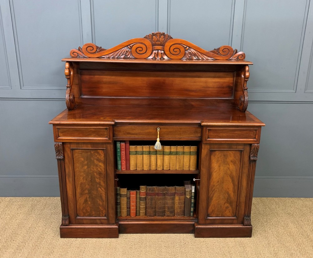 An early nineteenth-century flame mahogany chiffonier featuring a carved ornamental cresting, open central bookshelves, and two side cupboards. View XXI