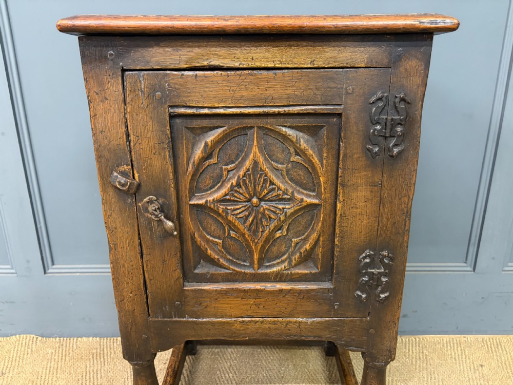 A small dark brown oak cupboard featuring a carved medallion door panel, wrought iron hardware, and turned legs. View V