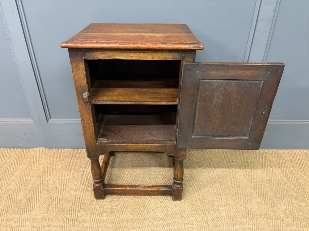 A small dark brown oak cupboard featuring a carved medallion door panel, wrought iron hardware, and turned legs. View VI