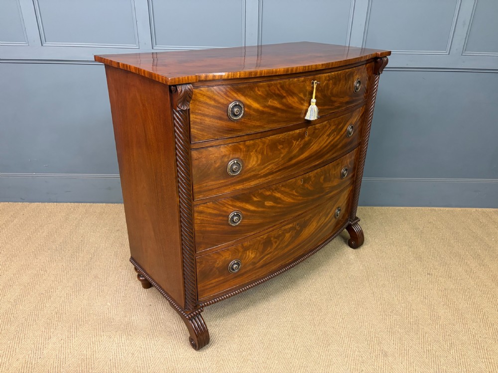 A Regency period mahogany bowfront chest of drawers featuring four graduated drawers, reeded side columns, brass ring handles, and turned feet.