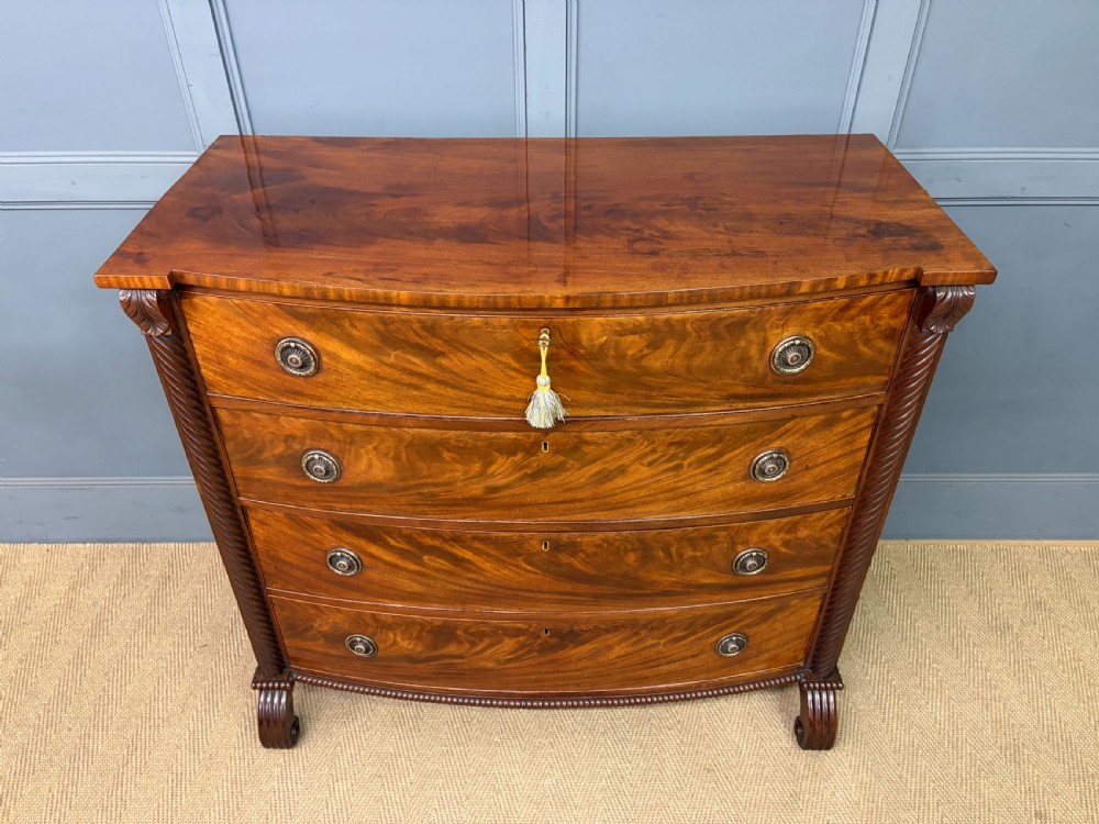 A Regency period mahogany bowfront chest of drawers featuring four graduated drawers, reeded side columns, brass ring handles, and turned feet. View III