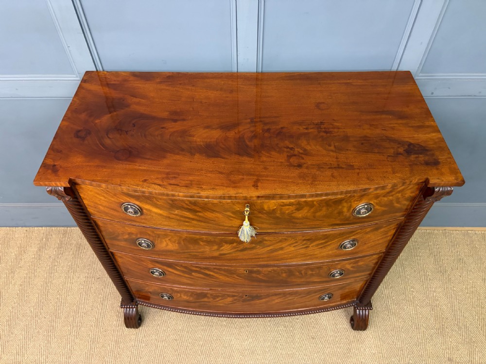 A Regency period mahogany bowfront chest of drawers featuring four graduated drawers, reeded side columns, brass ring handles, and turned feet. View IV