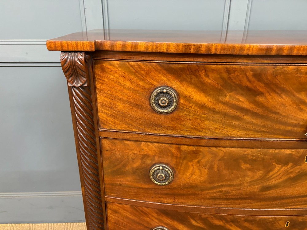 A Regency period mahogany bowfront chest of drawers featuring four graduated drawers, reeded side columns, brass ring handles, and turned feet. View V