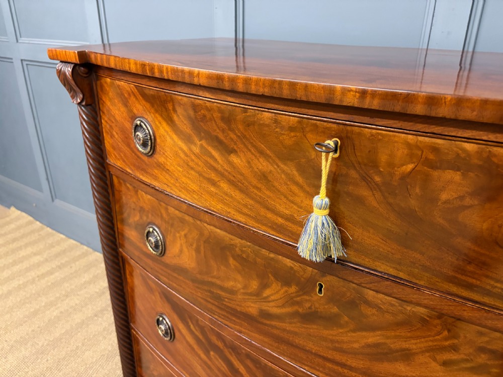 A Regency period mahogany bowfront chest of drawers featuring four graduated drawers, reeded side columns, brass ring handles, and turned feet. View VI