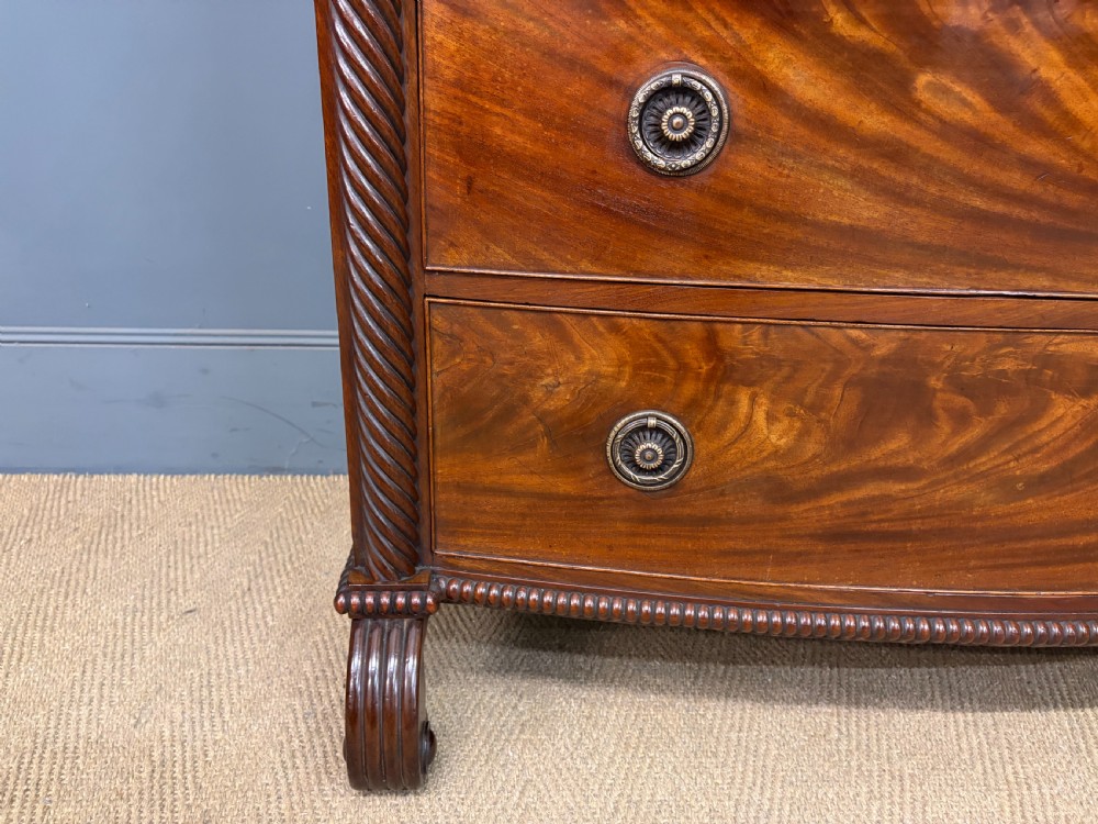 A Regency period mahogany bowfront chest of drawers featuring four graduated drawers, reeded side columns, brass ring handles, and turned feet. View IX