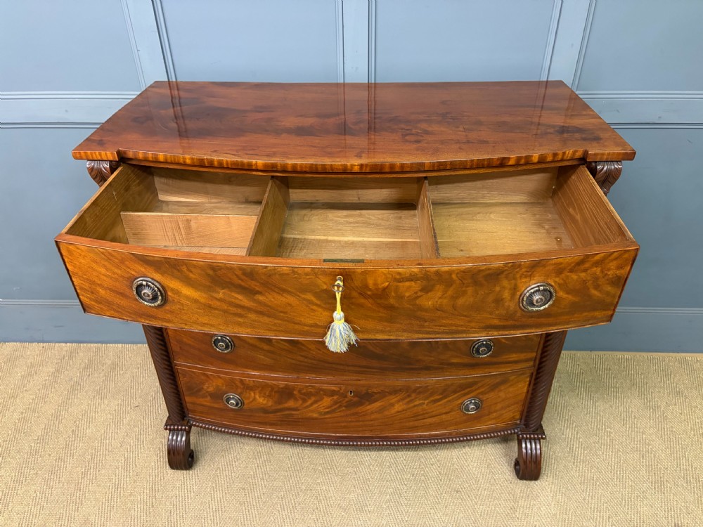 A Regency period mahogany bowfront chest of drawers featuring four graduated drawers, reeded side columns, brass ring handles, and turned feet. View XI