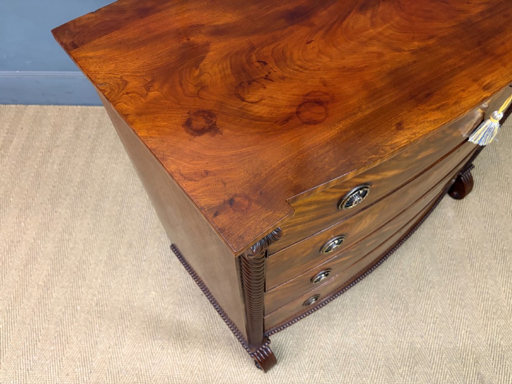 A Regency period mahogany bowfront chest of drawers featuring four graduated drawers, reeded side columns, brass ring handles, and turned feet. View XII