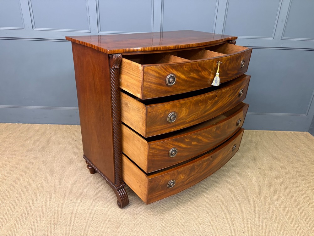 A Regency period mahogany bowfront chest of drawers featuring four graduated drawers, reeded side columns, brass ring handles, and turned feet. View XV