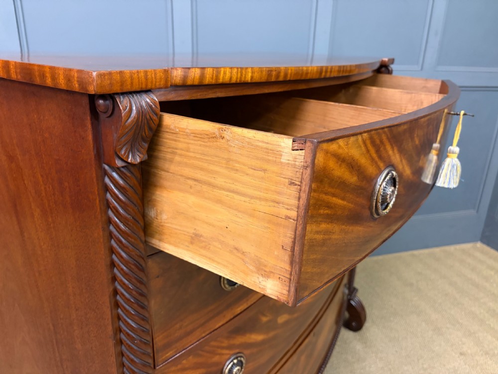 A Regency period mahogany bowfront chest of drawers featuring four graduated drawers, reeded side columns, brass ring handles, and turned feet. View XVI