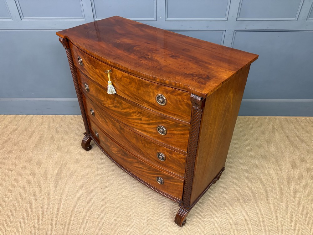 A Regency period mahogany bowfront chest of drawers featuring four graduated drawers, reeded side columns, brass ring handles, and turned feet. View XVII