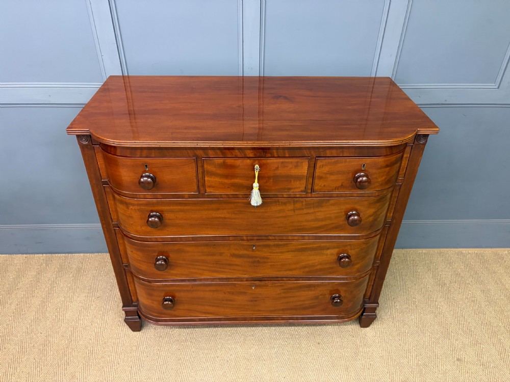 A George IV flame mahogany chest of drawers featuring a bowfront profile, turned wooden handles, and carved side columns. View IV