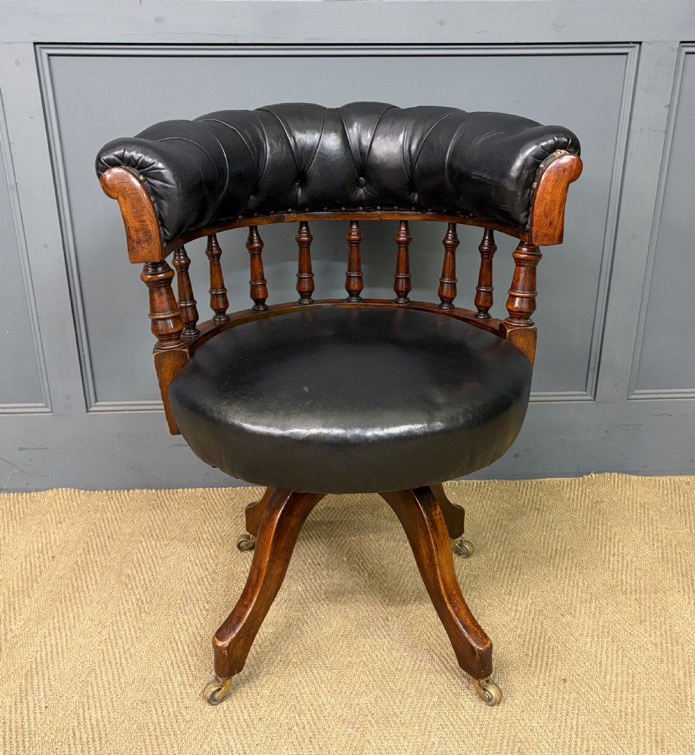 Victorian mahogany revolving desk chair featuring a deep button-tufted black leather curved back, turned spindles, a circular leather seat, and a swivel quadruped base with brass castors.