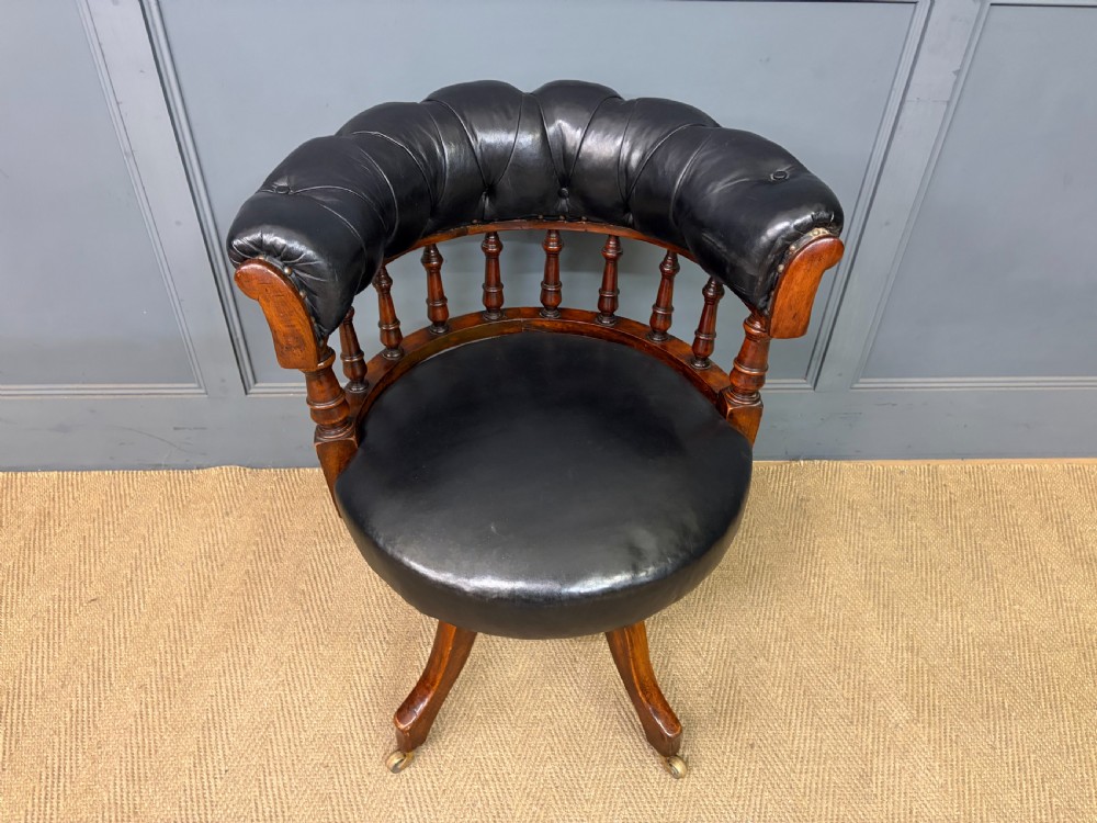 Victorian mahogany revolving desk chair featuring a deep button-tufted black leather curved back, turned spindles, a circular leather seat, and a swivel quadruped base with brass castors. View IV