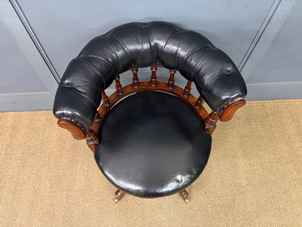Victorian mahogany revolving desk chair featuring a deep button-tufted black leather curved back, turned spindles, a circular leather seat, and a swivel quadruped base with brass castors. View V