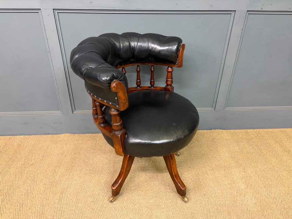 Victorian mahogany revolving desk chair featuring a deep button-tufted black leather curved back, turned spindles, a circular leather seat, and a swivel quadruped base with brass castors. View IX