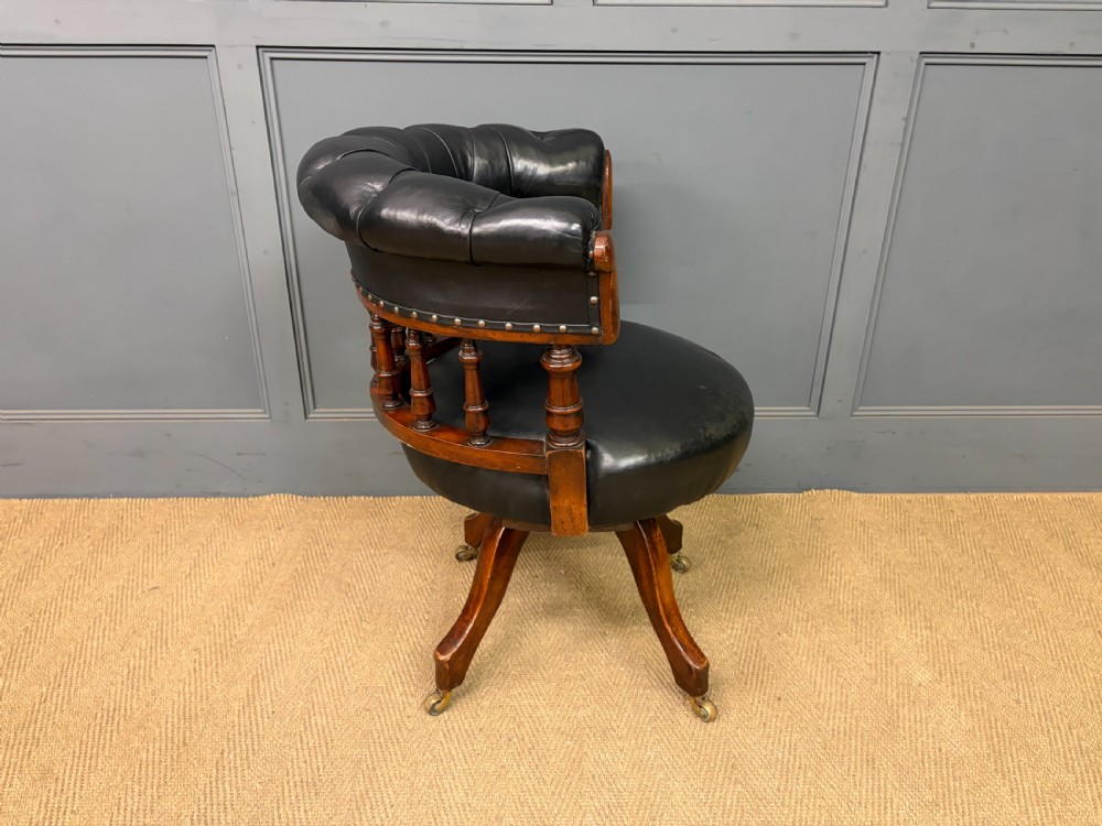 Victorian mahogany revolving desk chair featuring a deep button-tufted black leather curved back, turned spindles, a circular leather seat, and a swivel quadruped base with brass castors. View X