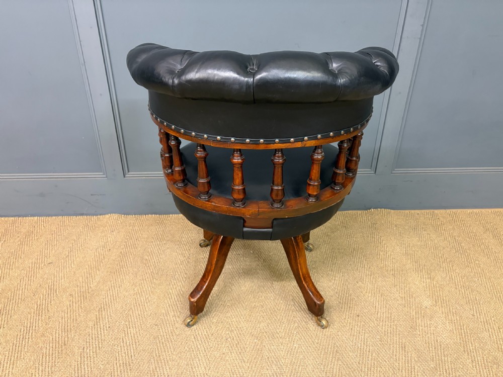 Victorian mahogany revolving desk chair featuring a deep button-tufted black leather curved back, turned spindles, a circular leather seat, and a swivel quadruped base with brass castors. View XI