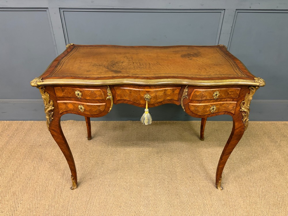 A 19th-century French Louis XV style writing desk with geometric parquetry veneers, gilt-bronze mounts, cabriole legs, and a warm brown leather writing surface.