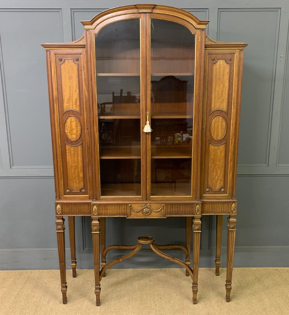 A Neoclassical mahogany display cabinet featuring a central glazed door, flanking inlaid wood cupboards, and tapered fluted legs connected by an ornate curved stretcher.