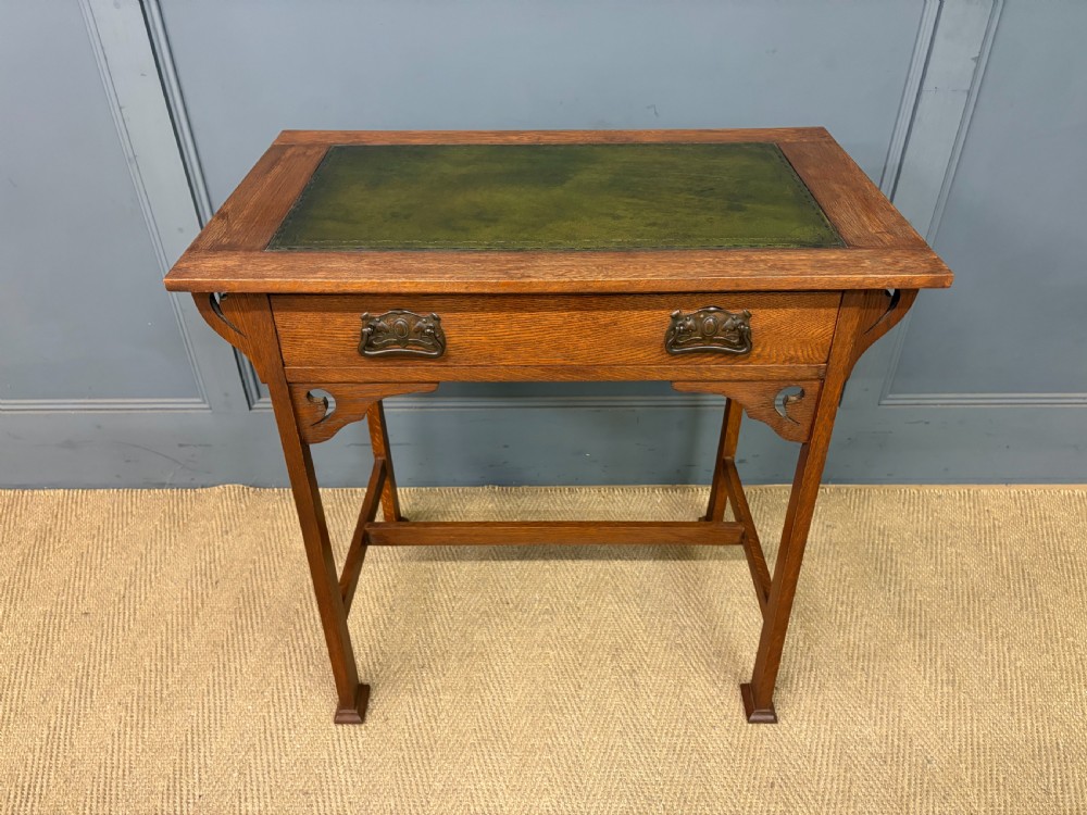 An Arts and Crafts period oak writing desk featuring an inset green leather top, single frieze drawer with brass handles, and pierced side supports.