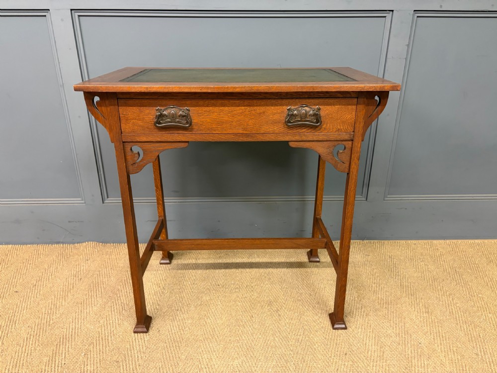 An Arts and Crafts period oak writing desk featuring an inset green leather top, single frieze drawer with brass handles, and pierced side supports. View III