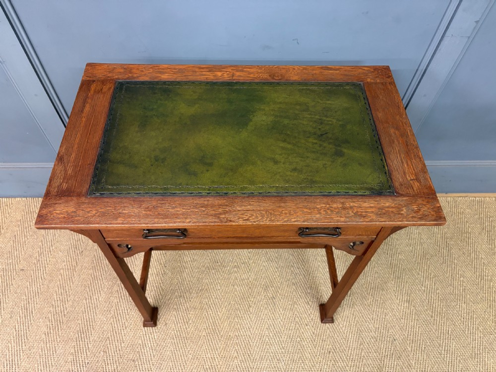 An Arts and Crafts period oak writing desk featuring an inset green leather top, single frieze drawer with brass handles, and pierced side supports. View IV