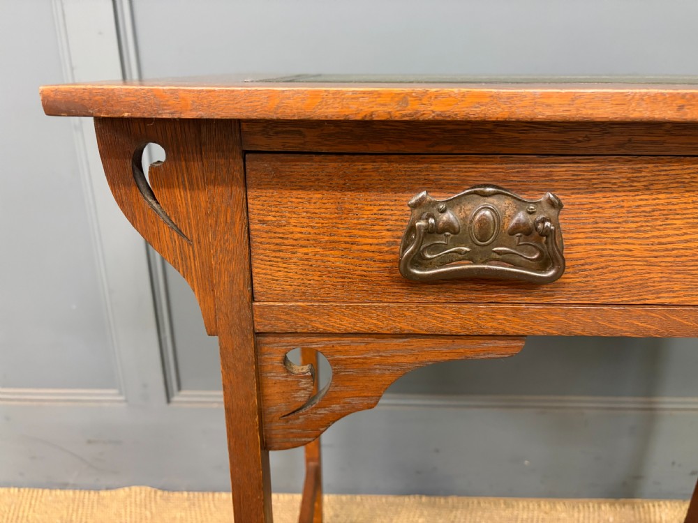 An Arts and Crafts period oak writing desk featuring an inset green leather top, single frieze drawer with brass handles, and pierced side supports. View V