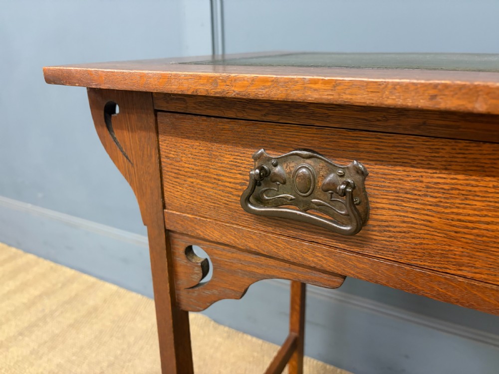 An Arts and Crafts period oak writing desk featuring an inset green leather top, single frieze drawer with brass handles, and pierced side supports. View VI