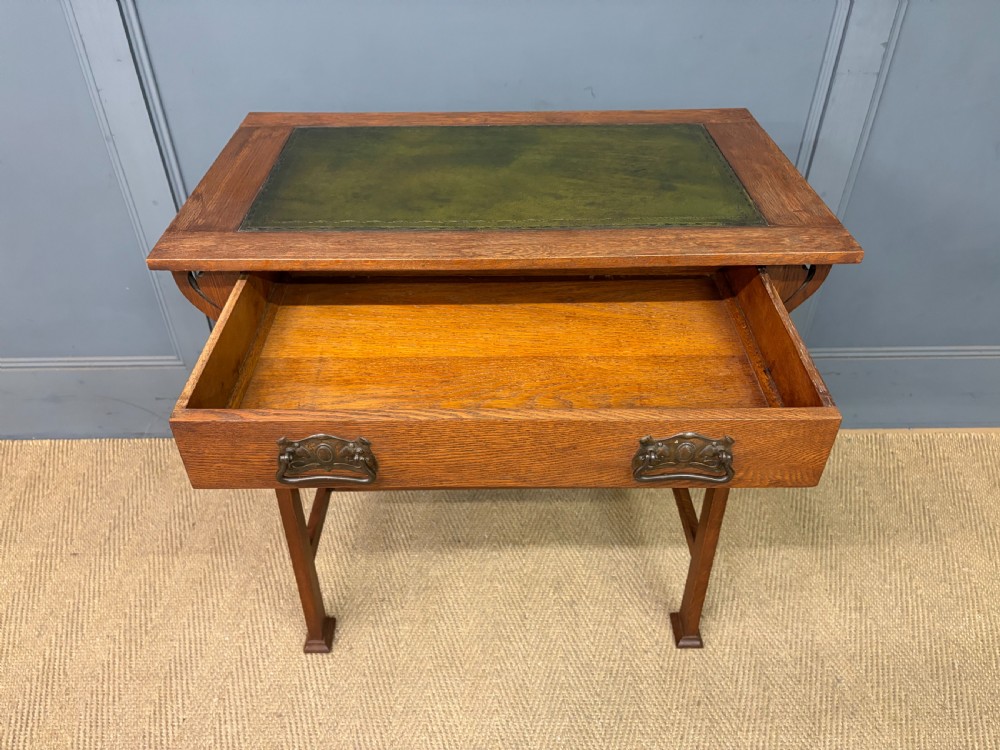 An Arts and Crafts period oak writing desk featuring an inset green leather top, single frieze drawer with brass handles, and pierced side supports. View VIII