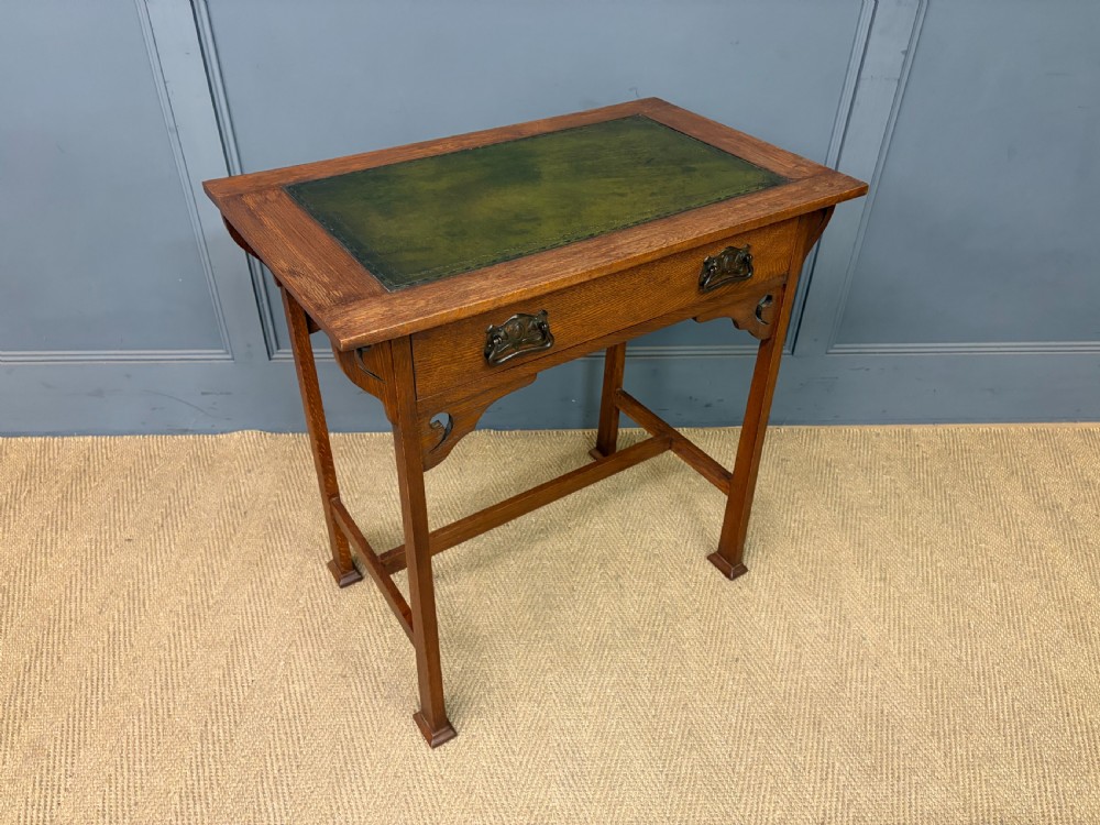 An Arts and Crafts period oak writing desk featuring an inset green leather top, single frieze drawer with brass handles, and pierced side supports. View IX