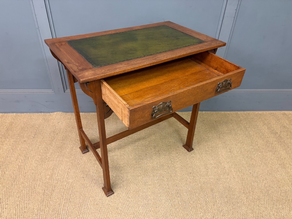 An Arts and Crafts period oak writing desk featuring an inset green leather top, single frieze drawer with brass handles, and pierced side supports. View X