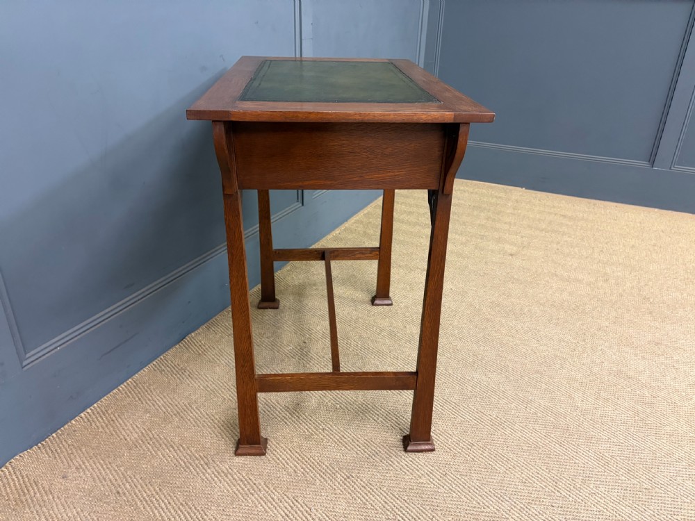 An Arts and Crafts period oak writing desk featuring an inset green leather top, single frieze drawer with brass handles, and pierced side supports. View XI
