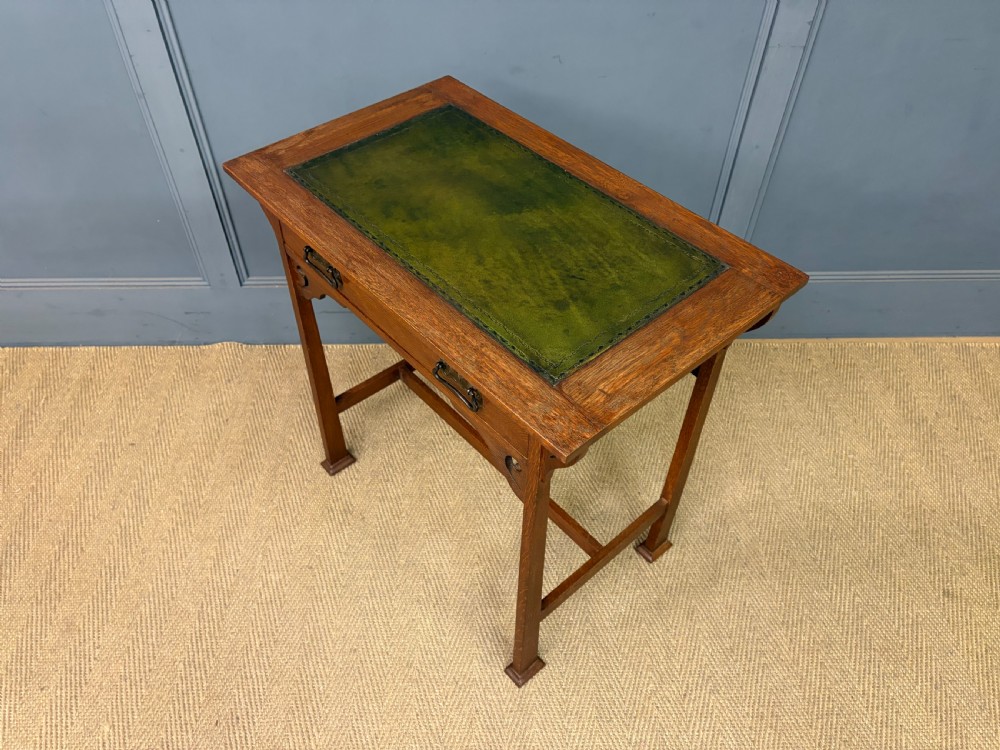 An Arts and Crafts period oak writing desk featuring an inset green leather top, single frieze drawer with brass handles, and pierced side supports. View XIII
