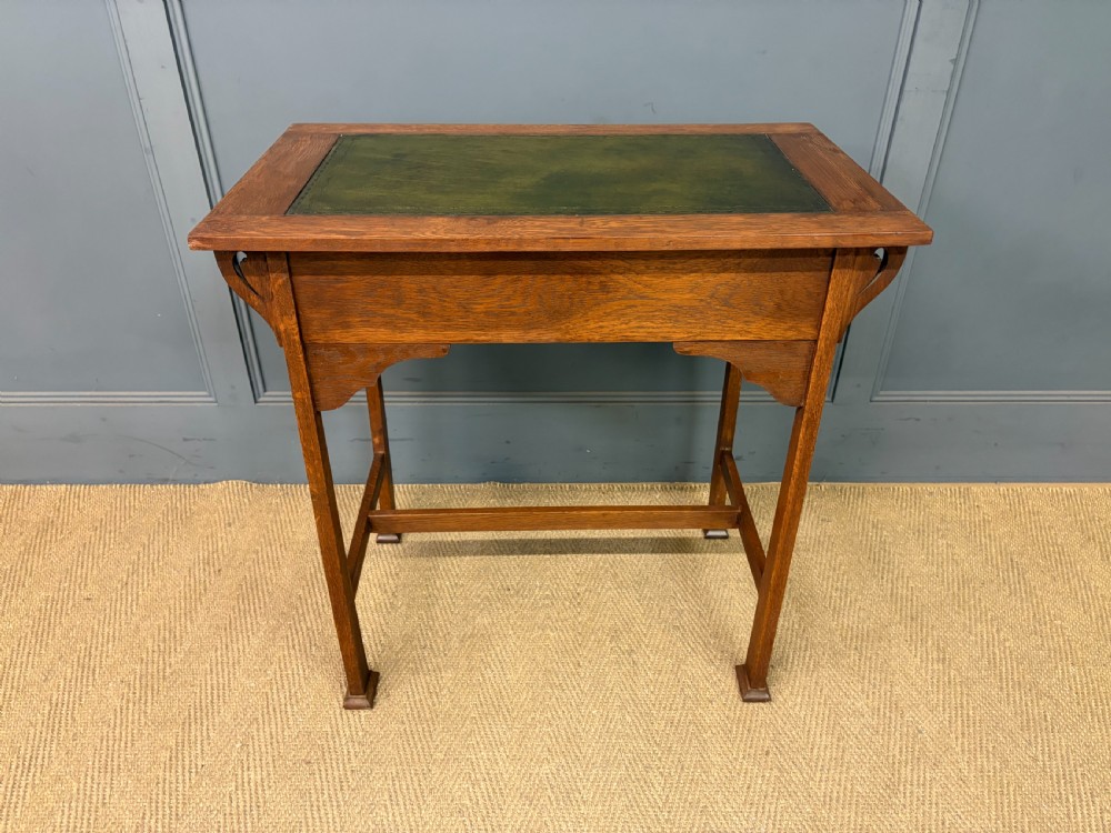 An Arts and Crafts period oak writing desk featuring an inset green leather top, single frieze drawer with brass handles, and pierced side supports. View XIV