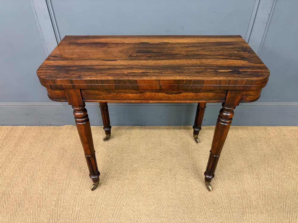 An early Victorian rosewood card table featuring a figured fold-over top, rectangular apron, turned tapered legs, and original brass castors.