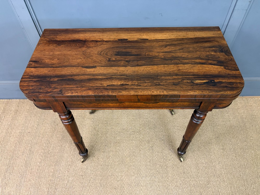 An early Victorian rosewood card table featuring a figured fold-over top, rectangular apron, turned tapered legs, and original brass castors. View IV