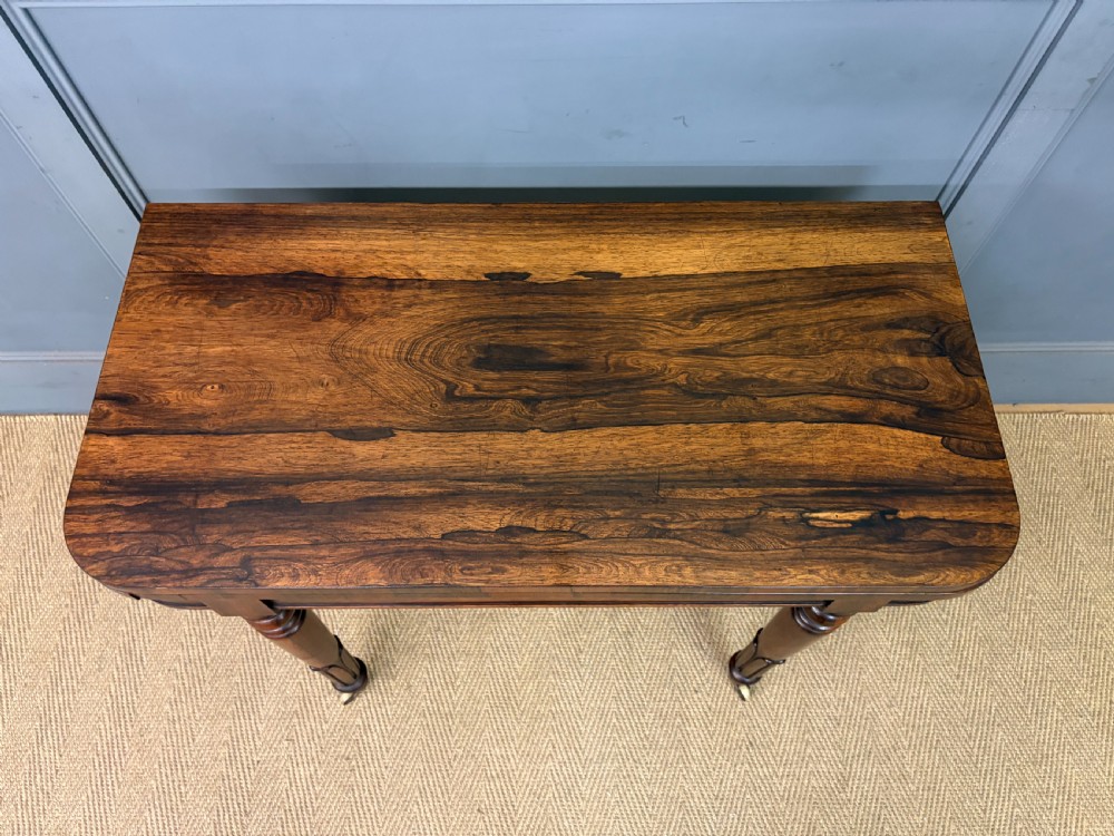 An early Victorian rosewood card table featuring a figured fold-over top, rectangular apron, turned tapered legs, and original brass castors. View V