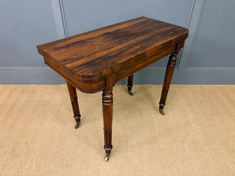 An early Victorian rosewood card table featuring a figured fold-over top, rectangular apron, turned tapered legs, and original brass castors. View X