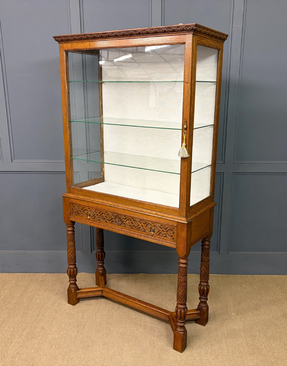 A tall carved oak display cabinet featuring three glass shelves, glazed sides, a carved frieze drawer, and carved turned legs.