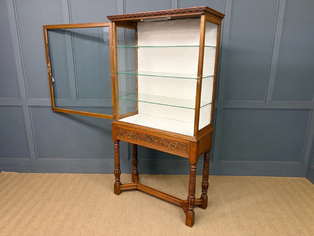 A tall carved oak display cabinet featuring three glass shelves, glazed sides, a carved frieze drawer, and carved turned legs. View III
