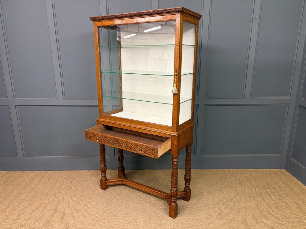 A tall carved oak display cabinet featuring three glass shelves, glazed sides, a carved frieze drawer, and carved turned legs. View IV