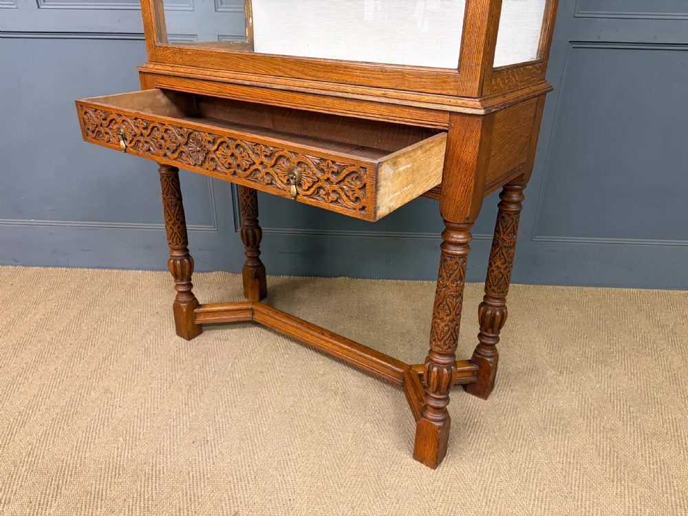 A tall carved oak display cabinet featuring three glass shelves, glazed sides, a carved frieze drawer, and carved turned legs. View V