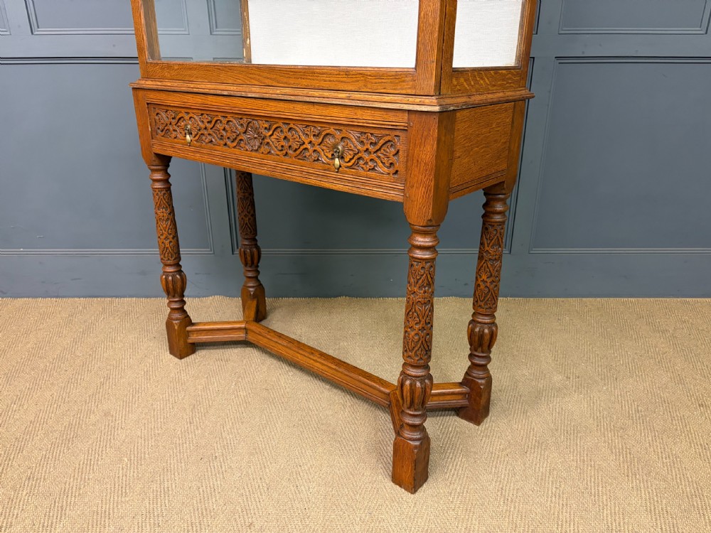 A tall carved oak display cabinet featuring three glass shelves, glazed sides, a carved frieze drawer, and carved turned legs. View VI