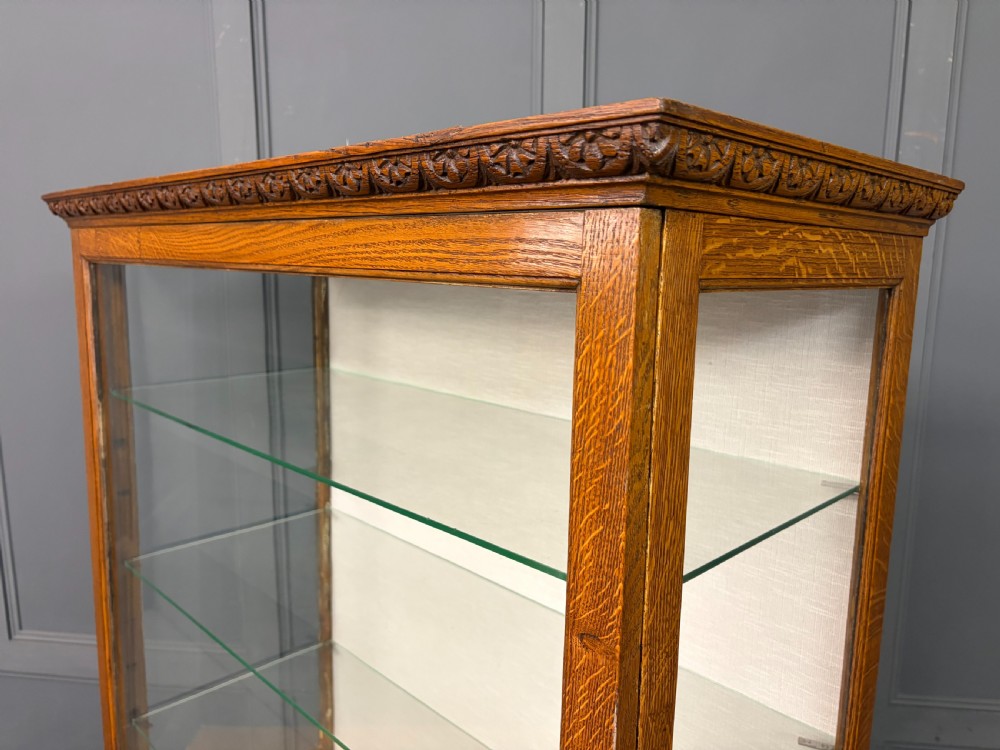 A tall carved oak display cabinet featuring three glass shelves, glazed sides, a carved frieze drawer, and carved turned legs. View VII