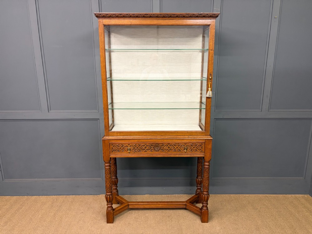 A tall carved oak display cabinet featuring three glass shelves, glazed sides, a carved frieze drawer, and carved turned legs. View VIII